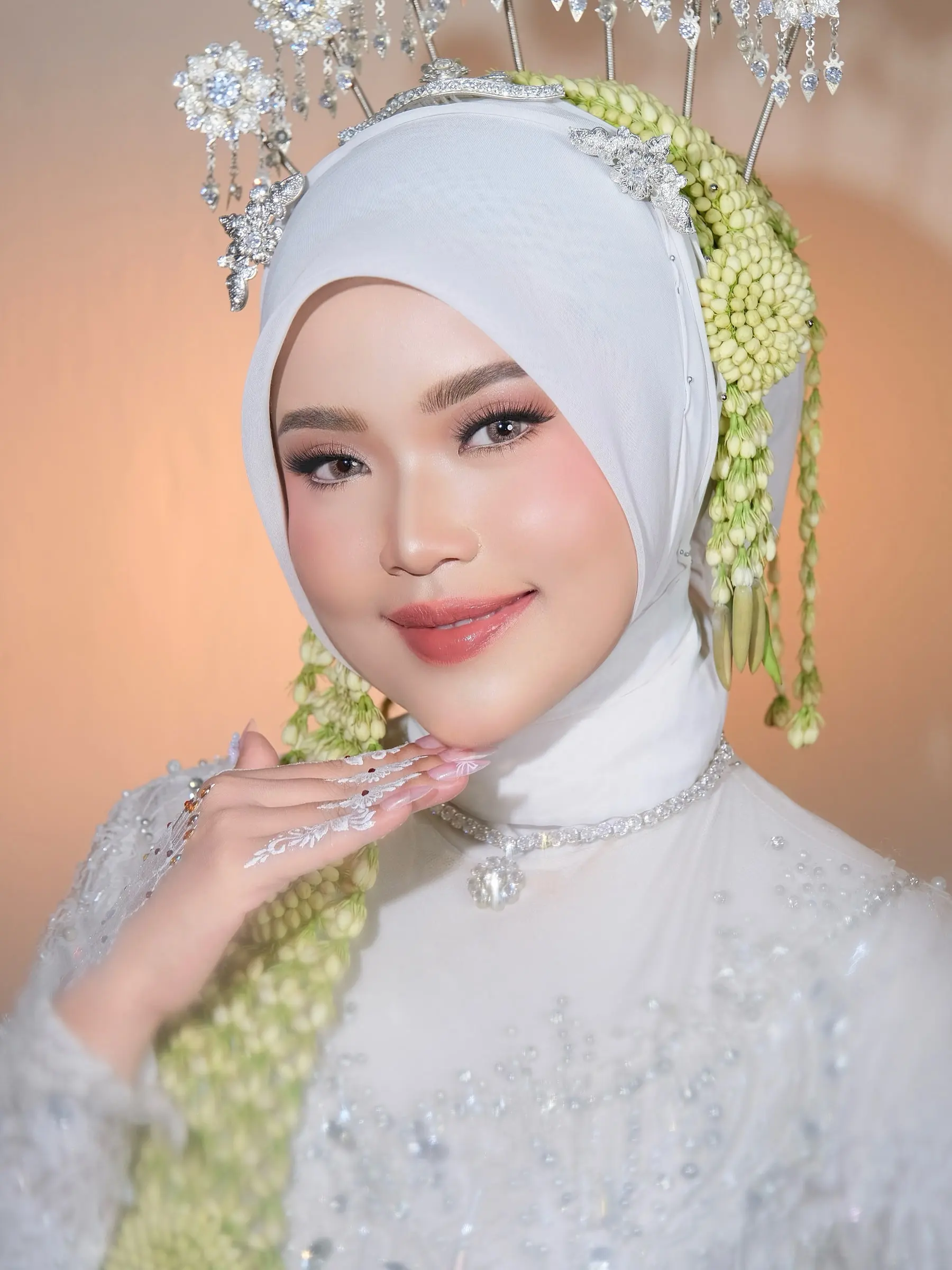 Riasan akad pengantin berhijab putih dengan siger perak dan ronce melati, berlatar warna persik