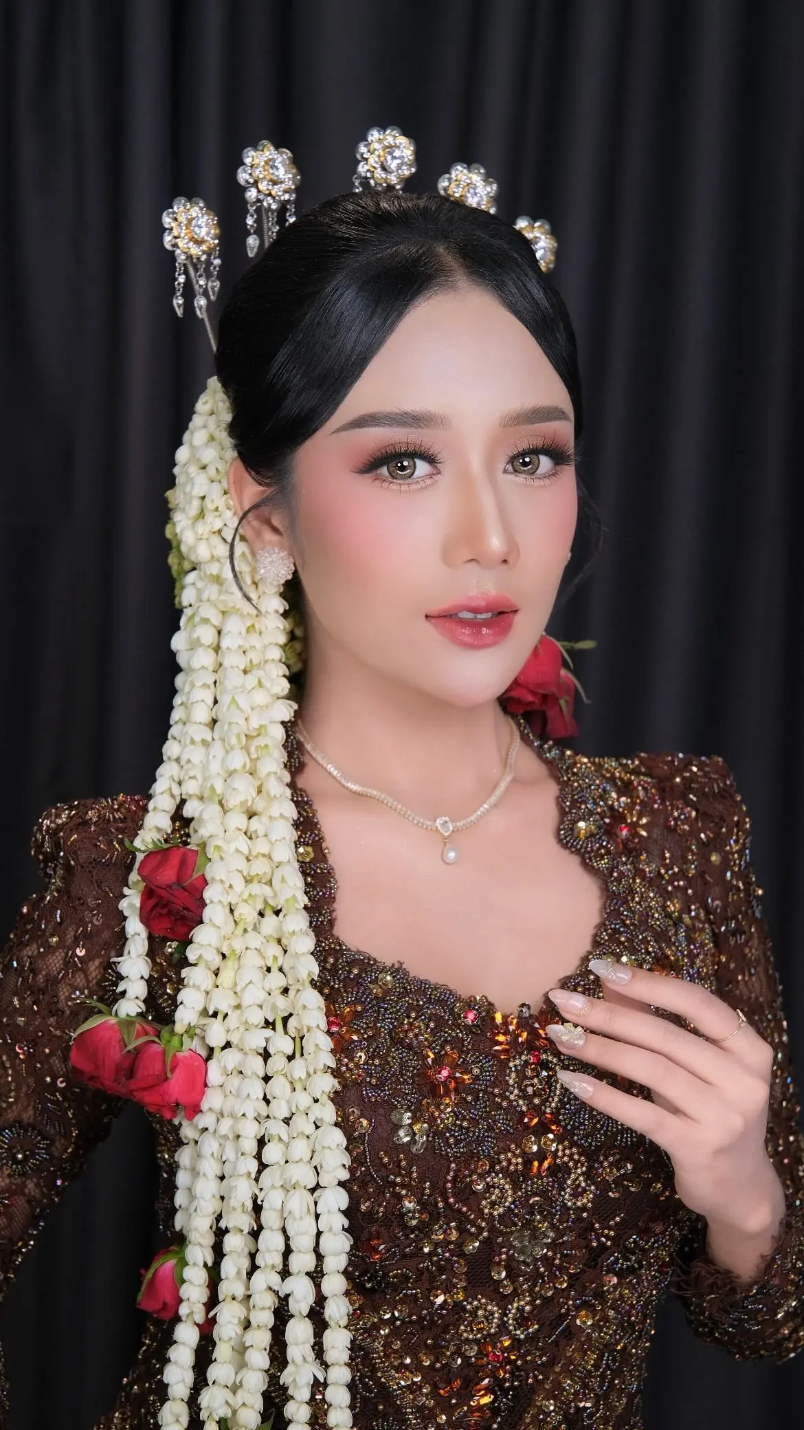 Riasan pengantin dengan untaian melati dan mawar merah, kebaya cokelat berpayet, berlatar gelap